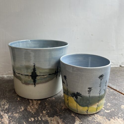 Two ceramic pots on a wooden windosill. The back ground is a white wall. The post show landscape with blue skies